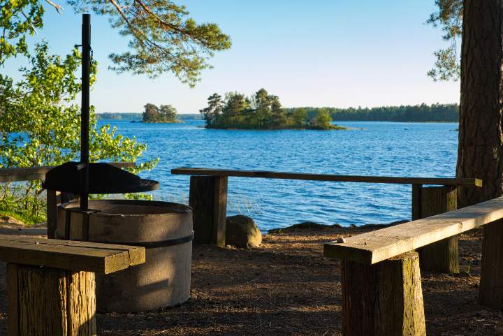 Ein Grillplatz mit einfachen Sitzbänken drum herum liegt direkt am Ufer des Asnen am Rand des Asnen Nationalpark. Kleine Inseln liegen im See und werden von der Abendsonne beschienen.