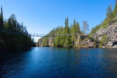Eine Fußgänger-Hängebrücke führt über einen Flusslauf, der sich in Richtung des Betrachters zu einem kleinen See erweitert. Die Ufer sind von steilen Felsen und Geröllfeldern gesäumt. Wo möglich wachsen hauptsächlich Nadelbäume. Die Szene liegt im nördlichen Teil des Hossa Nationalparks.