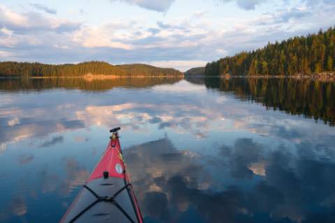 Die Spitze eines roten Kajaks im spiegelglatten Wasser des Kolovesi Nationalparks. Im Wasser spiegeln sich die Wolken und bewaldeten Inseln.