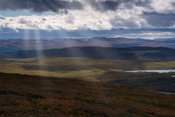 Sonnenstrahlen durchdringen die Wolken und hüllen die Landschaft des Pieljekaise Nationalparks in ein dramatisches Licht. Mehrere Bergketten staffeln sich hintereinander.