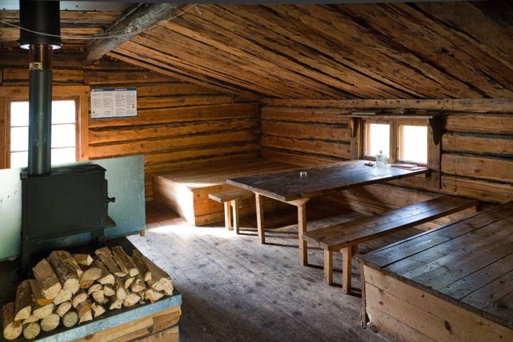 Im Inneren einer einfachen Hütte mit Sitz- und Liegegelegenheiten und einem einfachen Ofen, vor dem das Brennholz liegt.