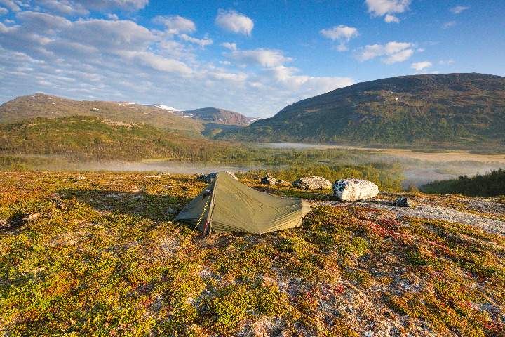 Ein kleines Zelt steht im Fjäll auf einer Kuppe mit wunderbarem Blick in den Vadvetjåkka Nationalpark. Im Flussdelta lösen sich die letzten Nebelschwaden auf.